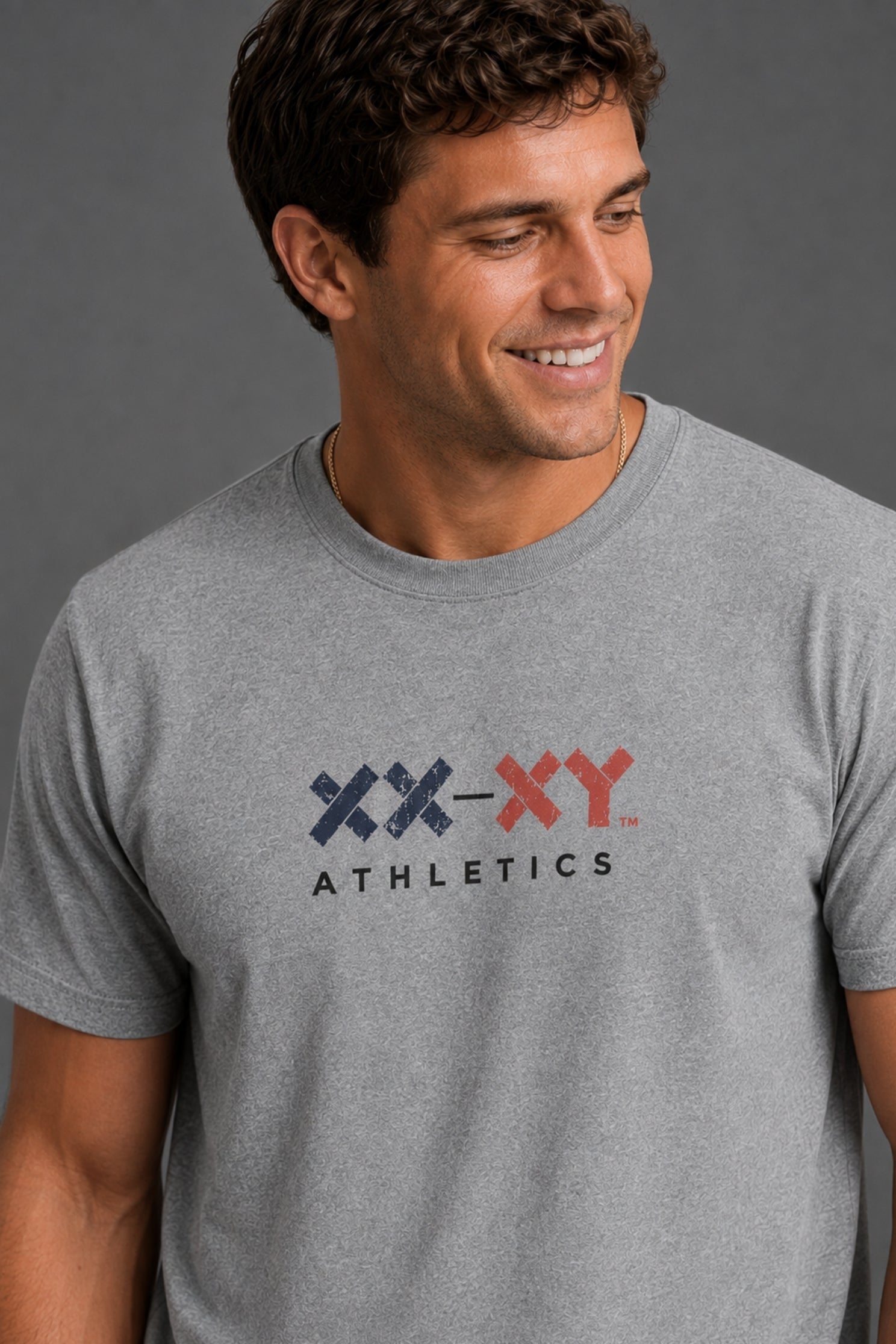 A man wearing a heather gray crewneck t-shirt with a 'XX-XY ATHLETICS' logo printed on the chest, set against a solid dark gray background. || Heather Gray