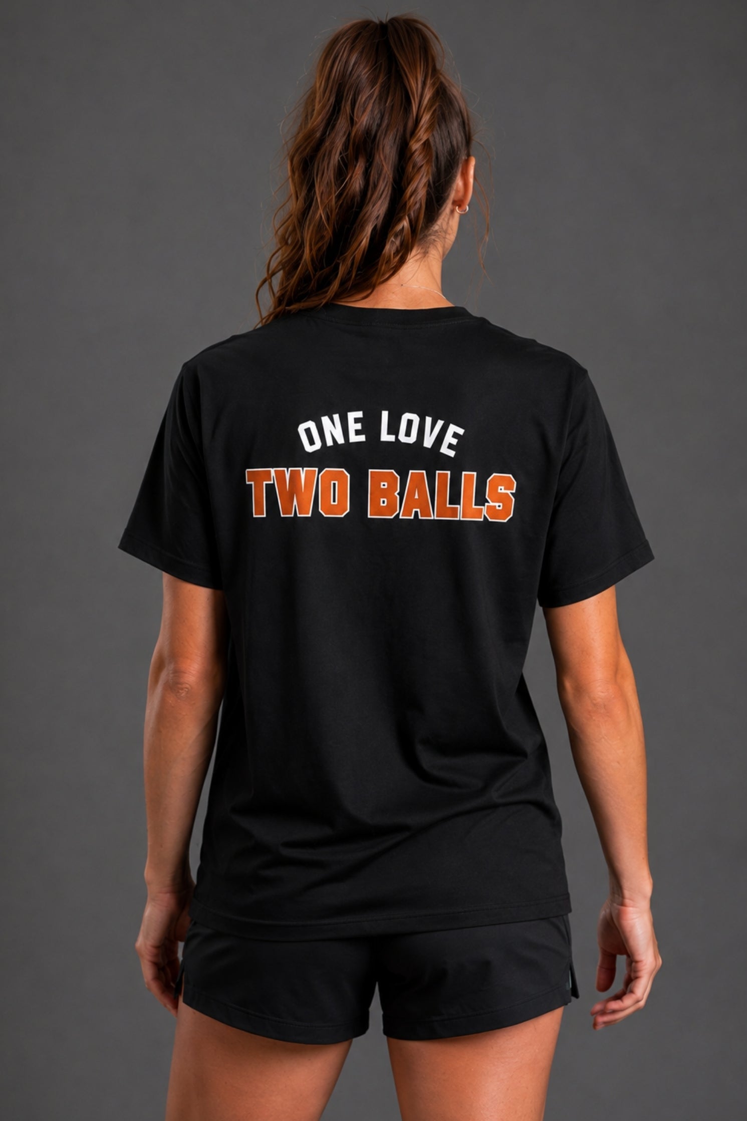 A woman shown from the back wearing a black short-sleeve t-shirt with the text 'ONE LOVE TWO BALLS' printed across the upper back in white and orange, paired with black athletic shorts against a dark gray studio background. || Black
