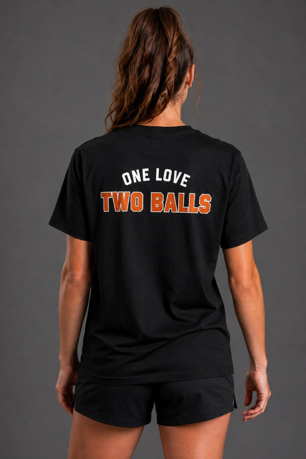 A woman shown from the back wearing a black short-sleeve t-shirt with the text 'ONE LOVE TWO BALLS' printed across the upper back in white and orange, paired with black athletic shorts against a dark gray studio background. || Black