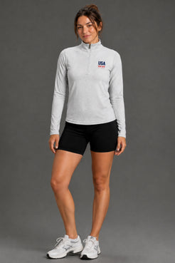 A woman modeling a light gray long-sleeve quarter-zip athletic pullover with a USA team logo on the left chest, paired with black athletic shorts and white sneakers, against a solid gray studio background. || Light Heather Gray - 1