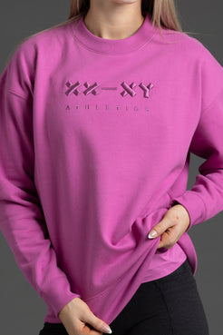 A close-up of a person wearing a bright pink crewneck sweatshirt with a tonal 'XX-XY ATHLETICS' logo printed across the chest, paired with dark gray leggings against a dark gray background. || Bright Fuchsia