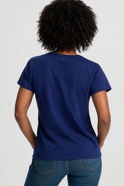 A woman with curly black hair wearing a navy blue short-sleeve t-shirt paired with blue jeans, against a light gray studio background. || Navy