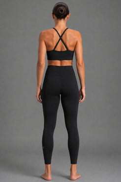 A woman viewed from the back wearing a black sports bra with crisscross straps and high-waisted black leggings, standing against a plain gray studio background. || Black Heather