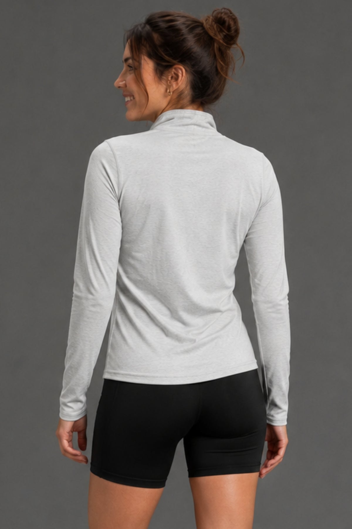A woman shown from the back wearing a light gray long-sleeve mock neck athletic top and black bike shorts against a solid gray studio background. || Light Heather Gray - 1