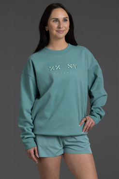 A woman modeling a matching teal sweatshirt and shorts set against a solid dark gray studio background. The sweatshirt features a tonal embroidered 'XX-SKY' logo on the chest. || Underseas