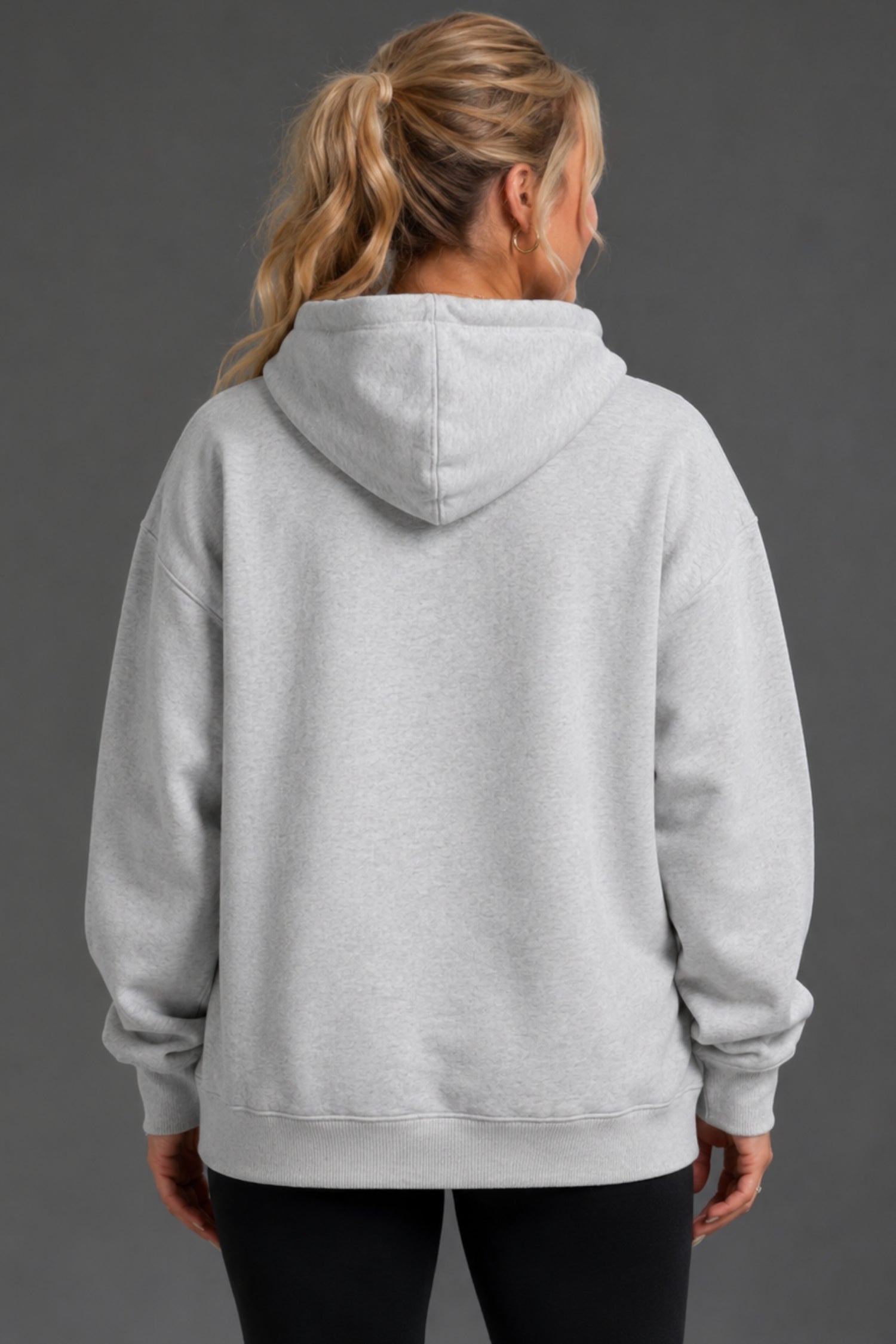 A woman viewed from the back wearing an oversized light gray heather pullover hoodie with black pants, set against a solid dark gray studio background. || Light Heather Gray