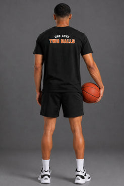 A man shown from behind wearing a black t-shirt with 'ONE LOVE TWO BALLS' printed in white and orange on the back, paired with black shorts, white socks, and sneakers, while holding a basketball against a gray studio background. || Black