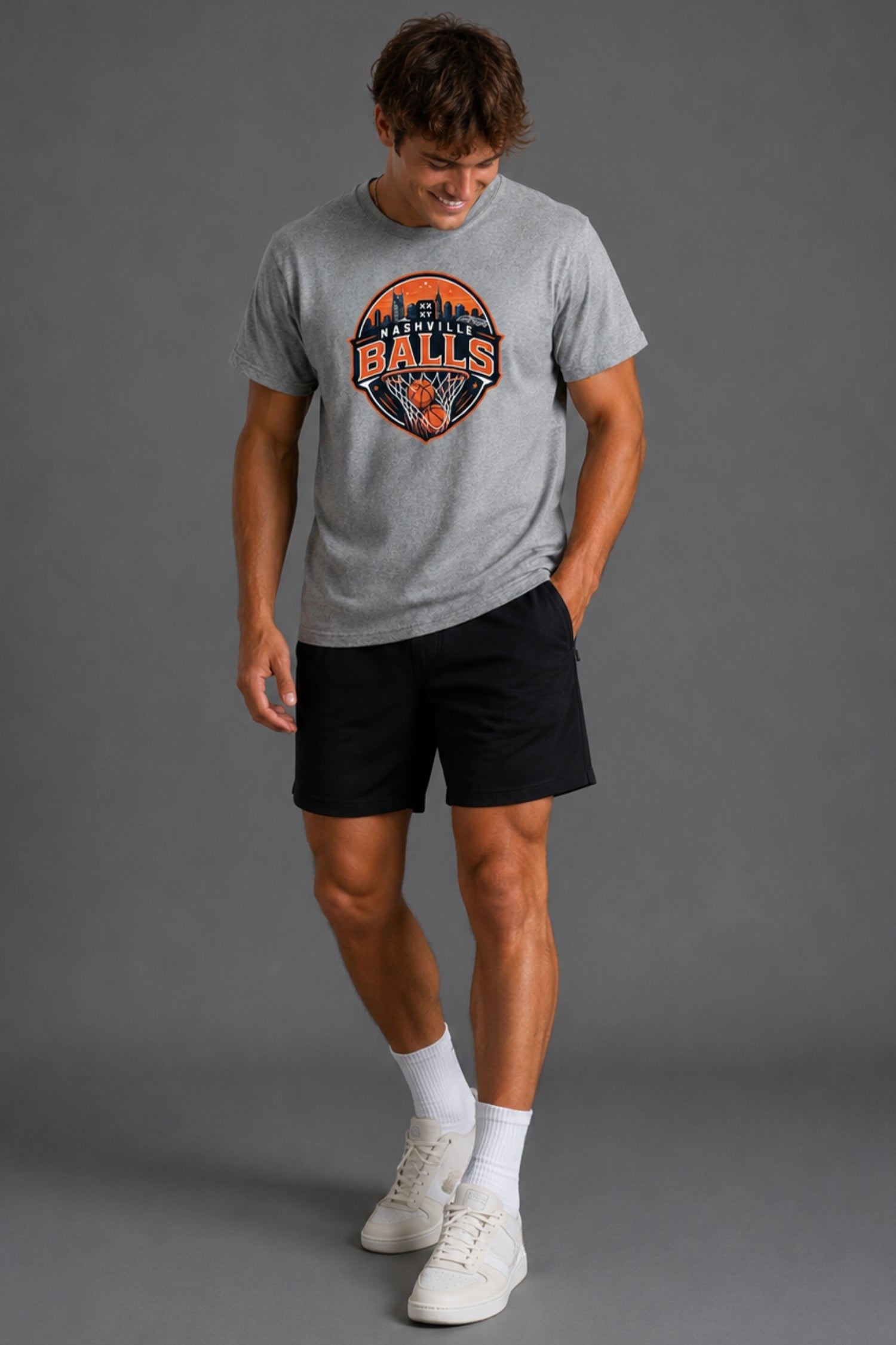 A man modeling a heather gray t-shirt with a 'Falcon Bats' graphic on the chest, paired with black athletic shorts, white socks, and white sneakers against a solid gray studio background. || Heather Gray