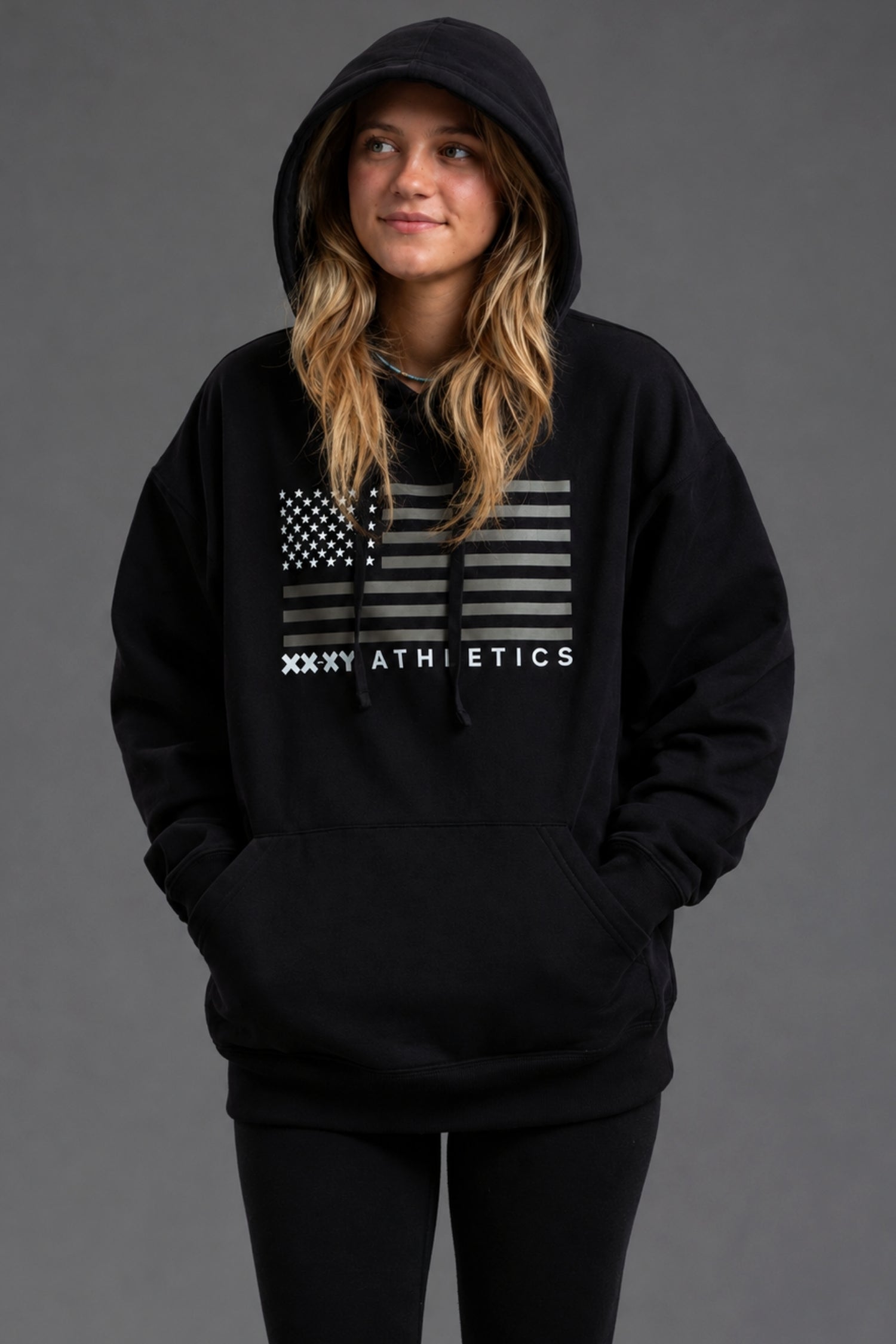 A woman wearing a black pullover hoodie with a distressed American flag graphic and 'XXXY ATHLETICS' text on the chest, posed against a solid dark gray studio background.