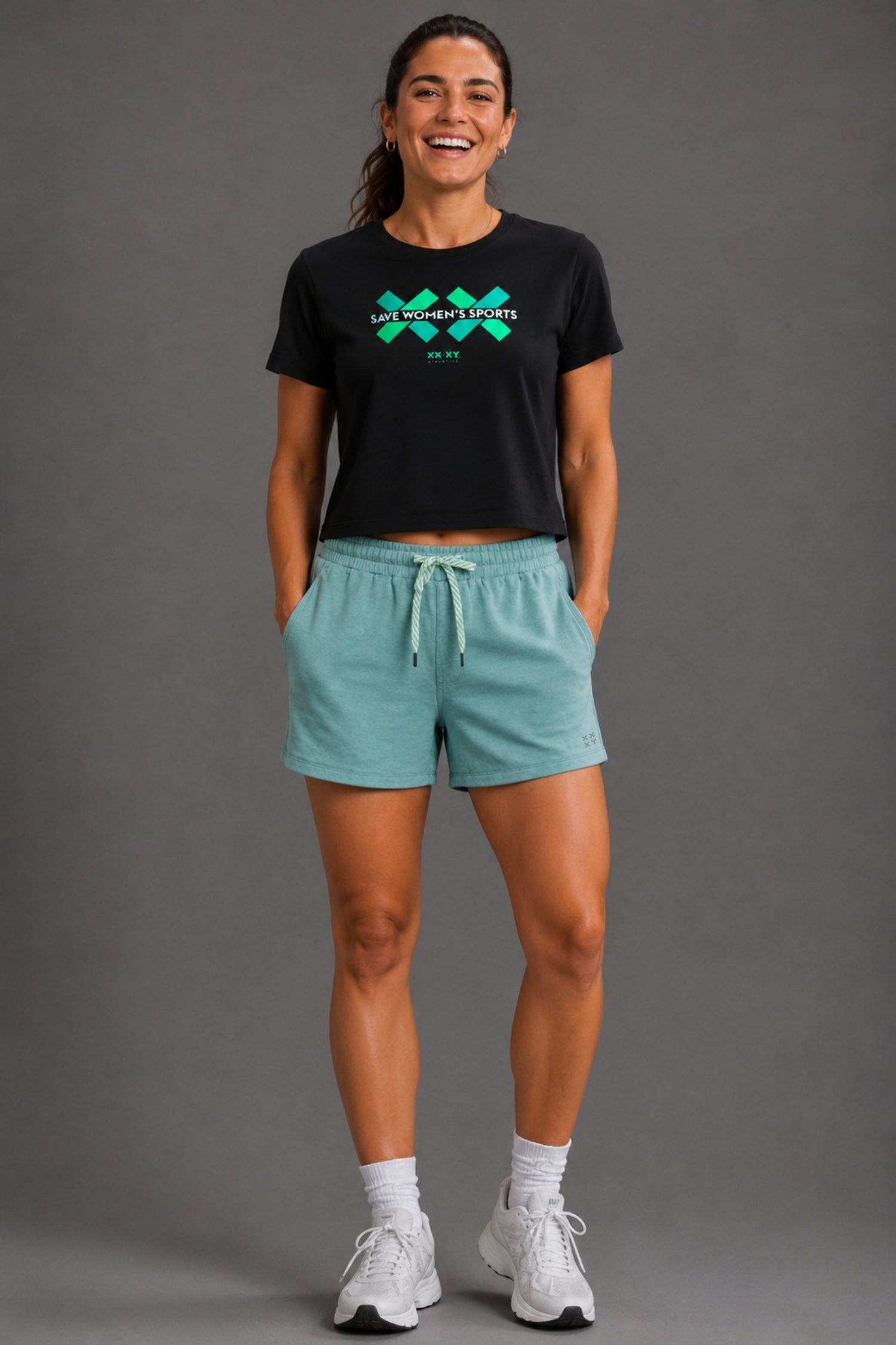 Woman wearing a black shirt with a "SAVE WOMEN'S SPORTS" text and green and blue graphics. Paired with blue shorts and white shoes on a gray background. || black