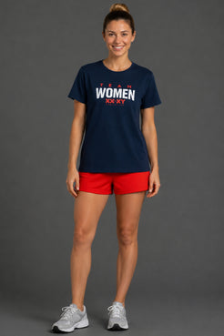 A woman modeling a navy blue t-shirt with 'TEAM WOMEN XXAT' printed in white and red on the chest, paired with red athletic shorts and white sneakers, against a solid gray studio background. || Navy
