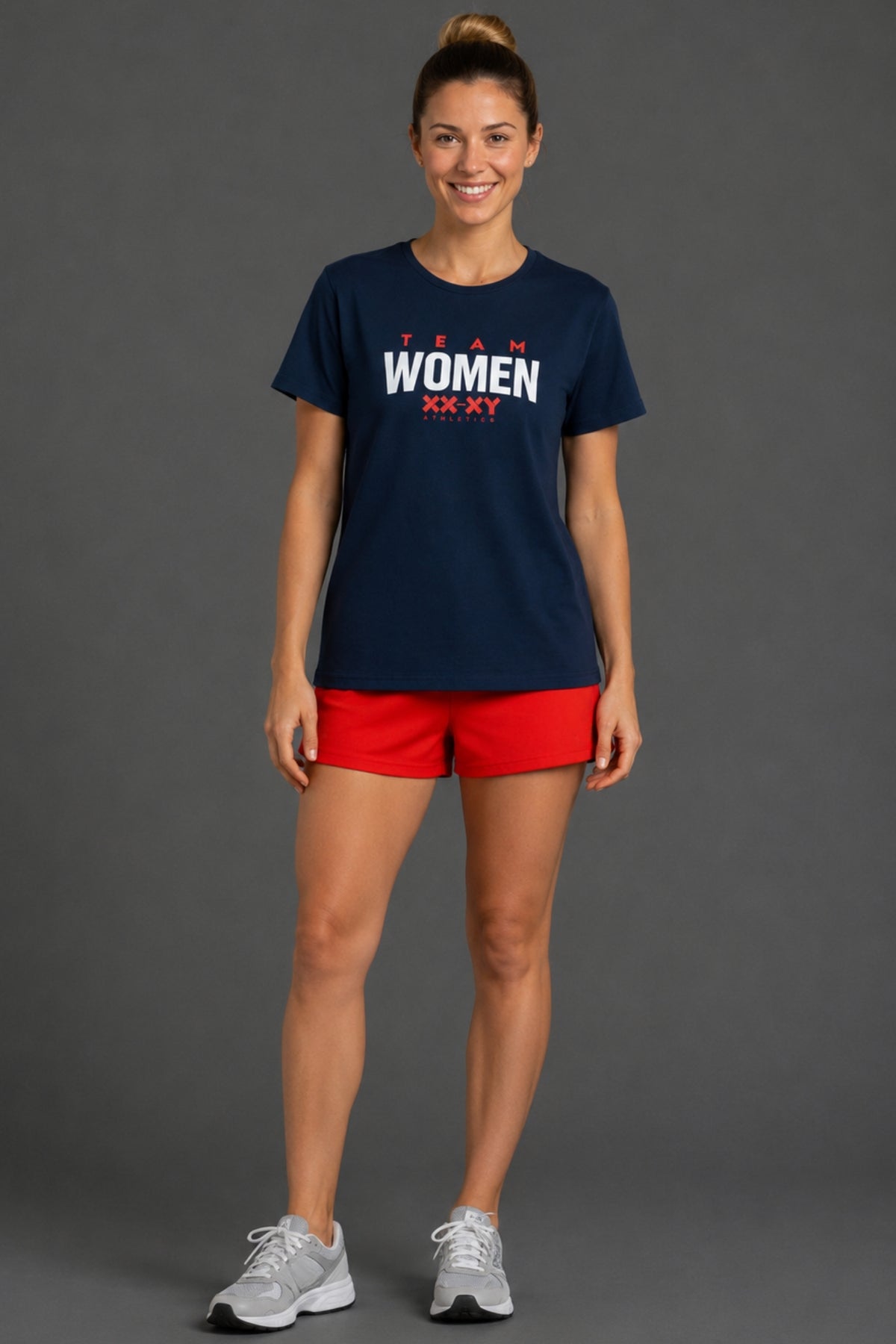 A woman modeling a navy blue t-shirt with 'TEAM WOMEN XXAT' printed in white and red on the chest, paired with red athletic shorts and white sneakers, against a solid gray studio background. || Navy