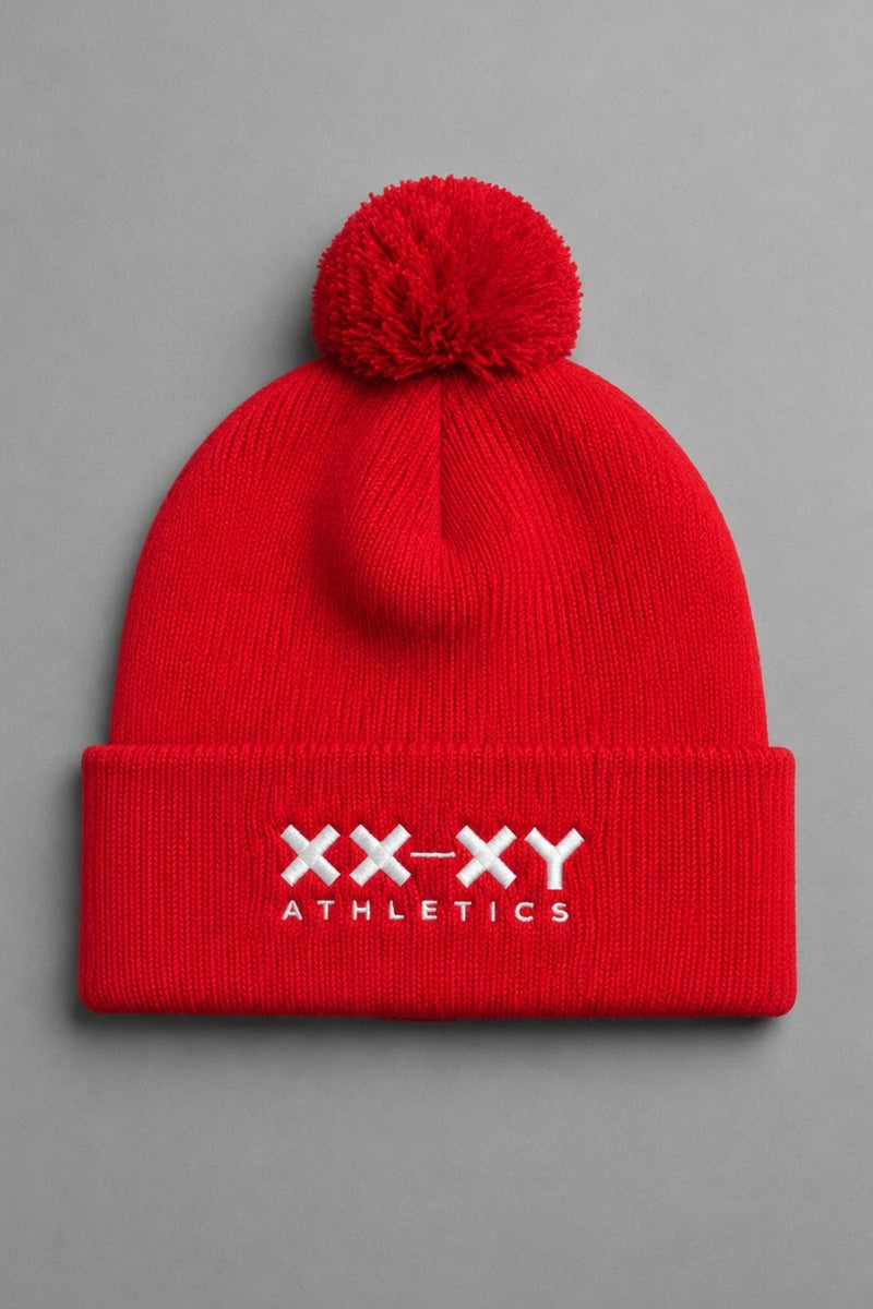 A bright red knit pom-pom beanie with a folded cuff featuring the white embroidered text 'XX-XY ATHLETICS', shown against a solid gray background. || Red