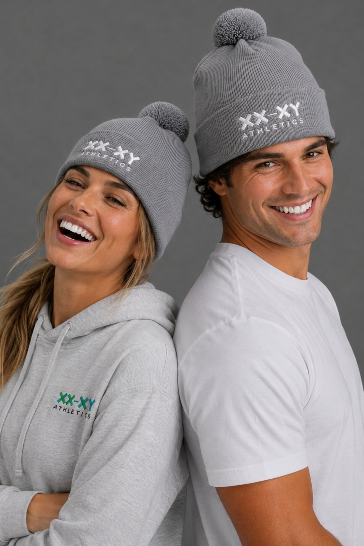 Male and female models wearing gray XX-XY Athletics pom beanies, a gray logo hoodie, and a white T-shirt, posing back-to-back against a gray studio background.