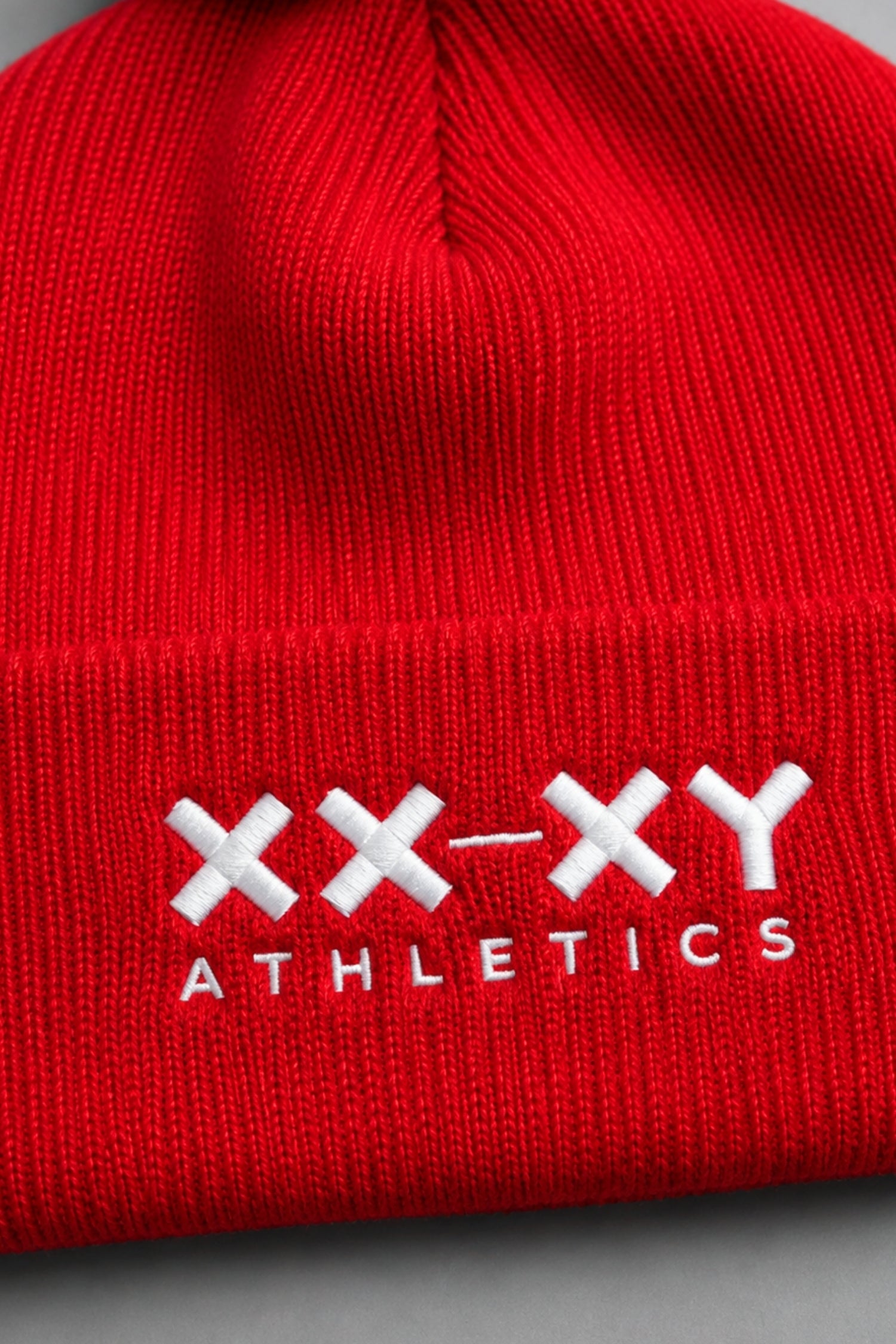 A close-up of a bright red ribbed knit beanie with white embroidered 'XX-XY ATHLETICS' branding on the front cuff. || Red