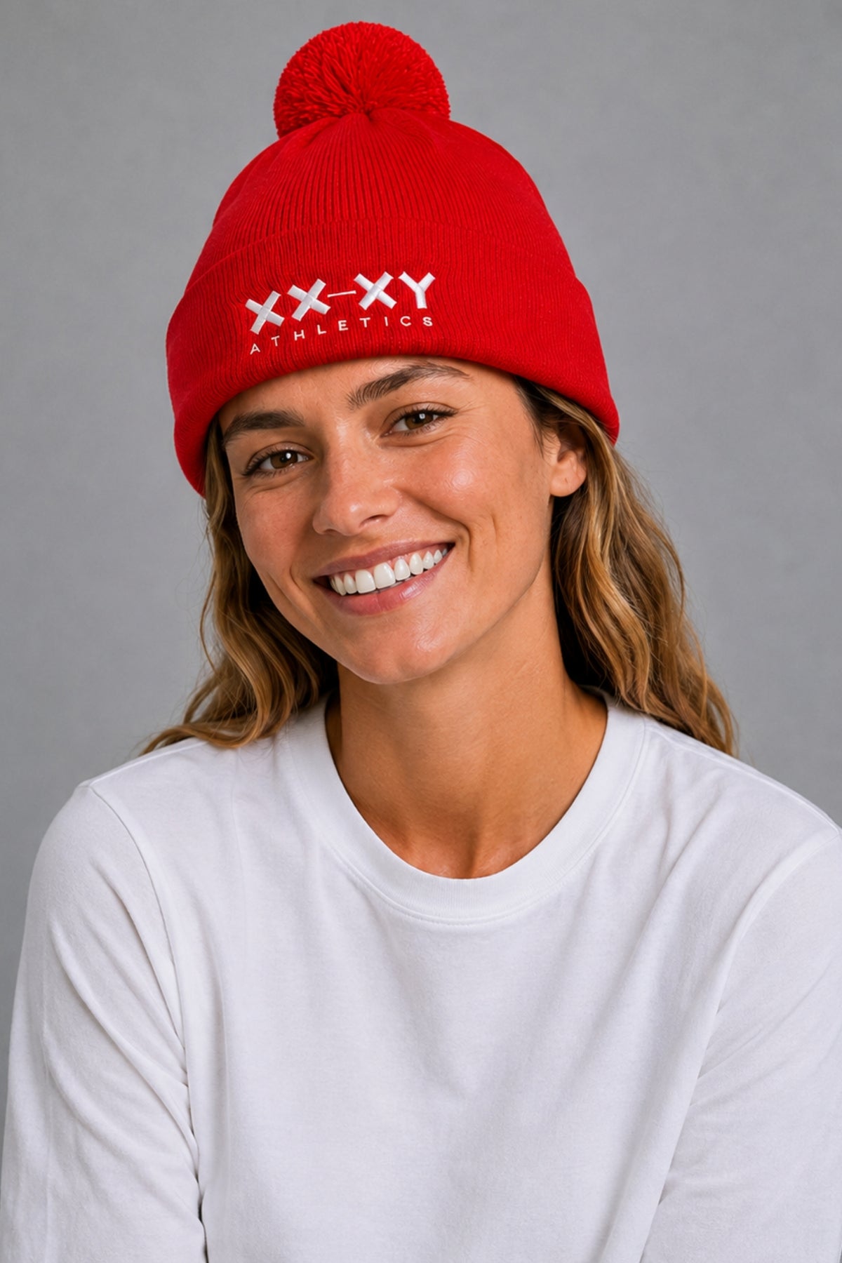 A smiling woman wearing a bright red knit beanie with a pom-pom and white embroidered 'XX-XY ATHLETICS' text on the front, paired with a white long-sleeve shirt against a plain gray studio background.