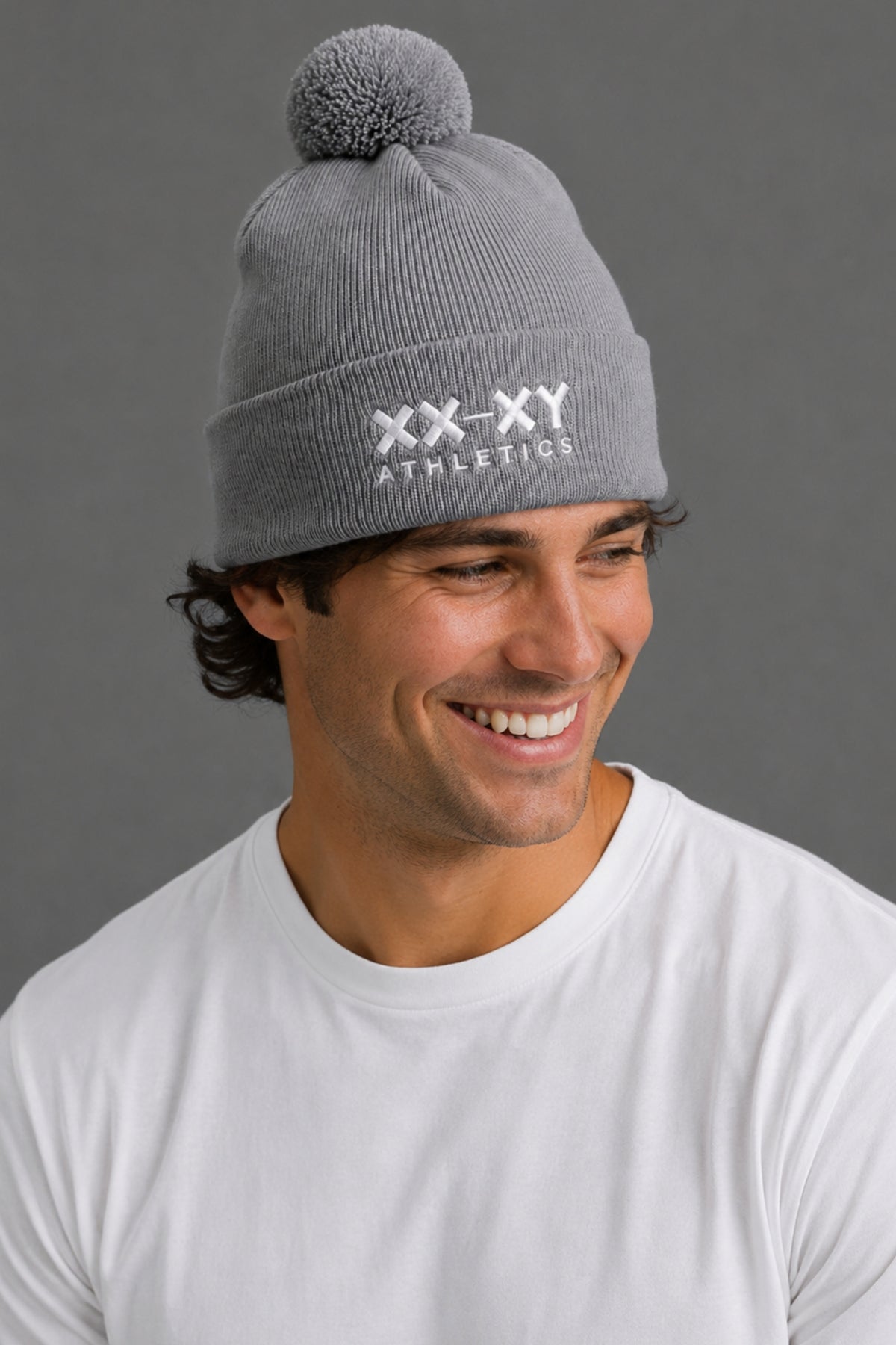A smiling man wearing a gray ribbed knit beanie with a pom-pom and white embroidered 'XX-XY ATHLETICS' text on the front, against a plain gray background.