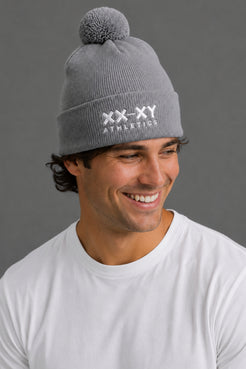 A smiling man wearing a gray ribbed knit beanie with a pom-pom and white embroidered 'XX-XY ATHLETICS' text on the front, against a plain gray background. || Heather Gray