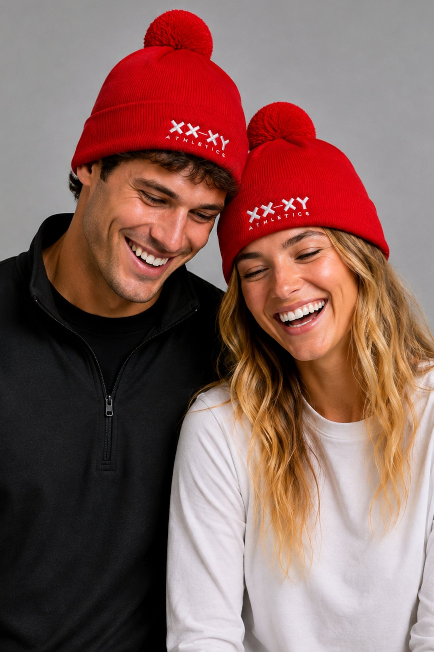A man and a woman wearing matching red knit pom-pom beanies with white 'XX-XY FANTASY' text on the front, posed against a plain gray studio background. || red
