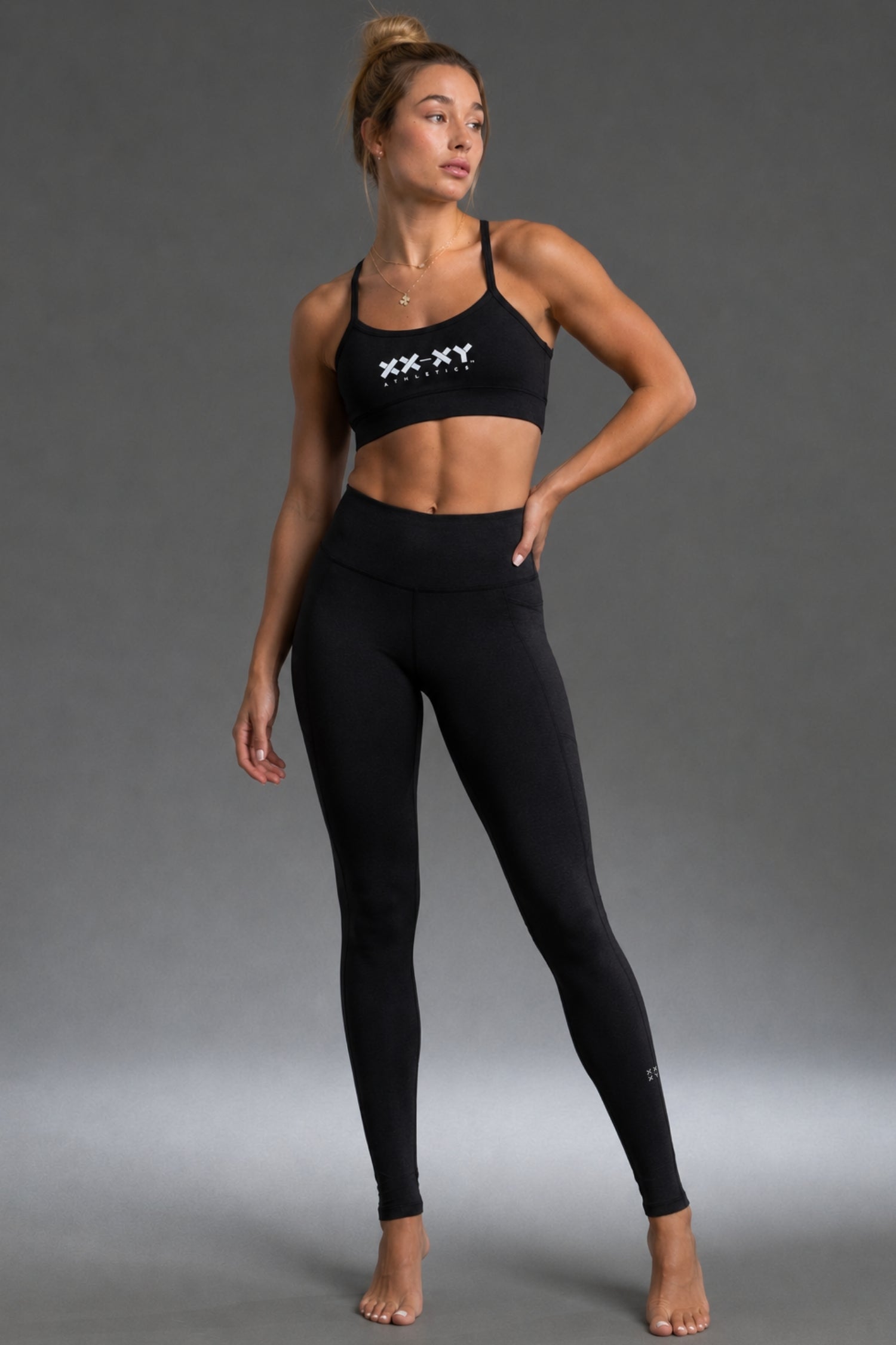 A woman modeling a black two-piece activewear set consisting of a sports bra with a white 'XX-XY' logo on the chest and matching high-waisted leggings, set against a solid gray studio background. || Black Heather