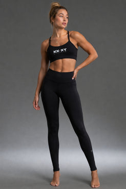 A woman modeling a black two-piece activewear set consisting of a sports bra with a white 'XX-XY' logo on the chest and matching high-waisted leggings, set against a solid gray studio background. || Black Heather