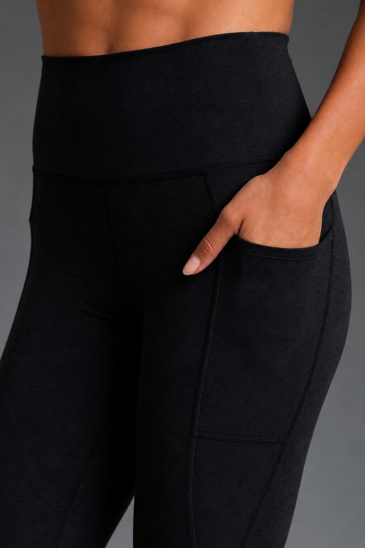 Close-up of a person wearing black high-waisted leggings with a side pocket, shown against a dark gray studio background. || Black Heather