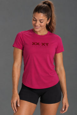 Female model wearing a fuchsia XX-XY Athletics T-shirt with black bike shorts against a gray studio background. || Berry