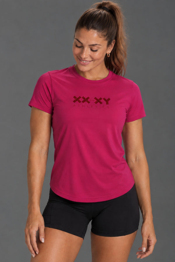 Female model wearing a fuchsia XX-XY Athletics T-shirt with black bike shorts against a gray studio background. || Berry