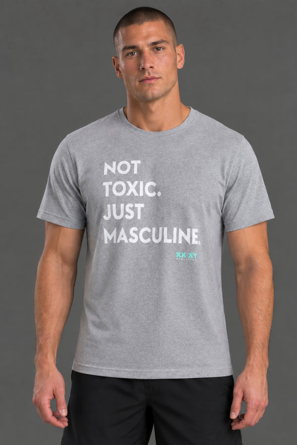 Male model wearing a gray “Not Toxic. Just Masculine.” graphic T-shirt with black shorts against a gray studio background. || Light Heather Gray