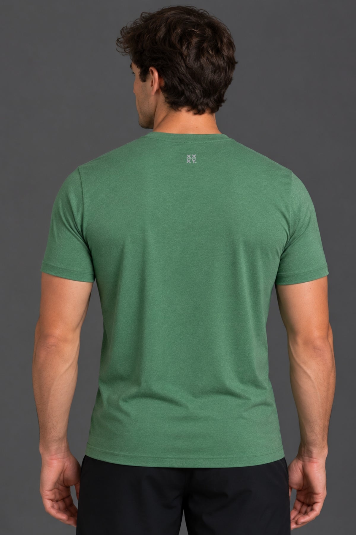 A man viewed from the back wearing a short-sleeve crew-neck t-shirt in a heathered green color, paired with black pants, against a solid dark gray studio background.