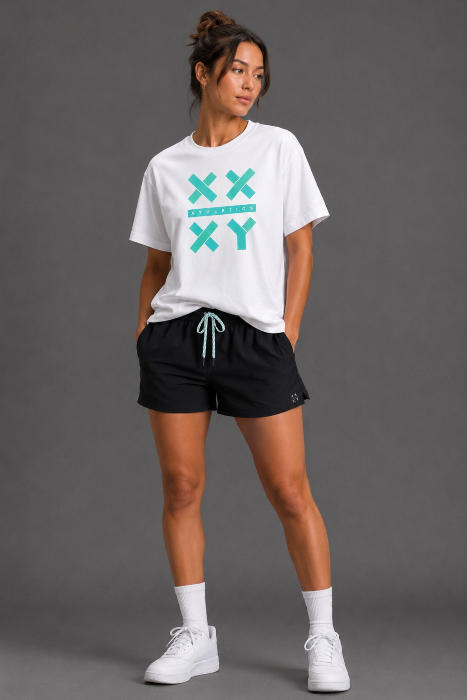 A woman modeling a white graphic t-shirt with a teal 'XX YY' design, paired with black athletic shorts, white socks, and white sneakers, against a solid gray studio background. || White