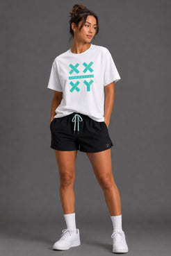 A woman modeling a white graphic t-shirt with a teal 'XX YY' design, paired with black athletic shorts, white socks, and white sneakers, against a solid gray studio background. || White