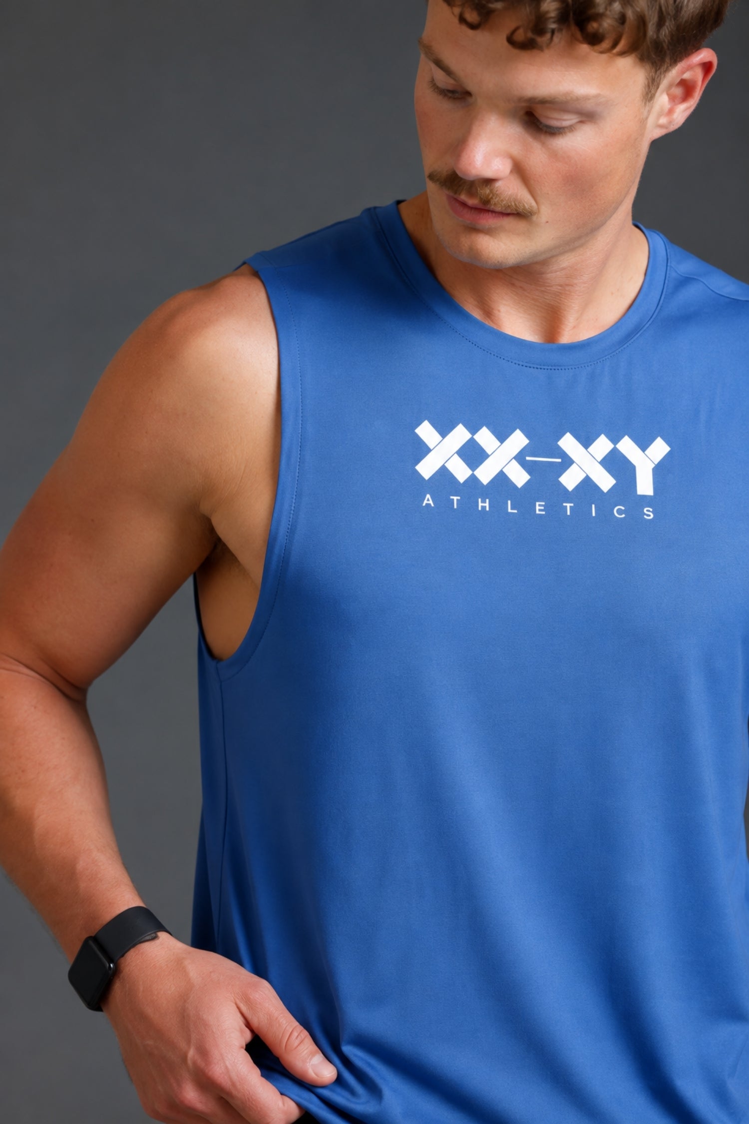 A man wearing a royal blue sleeveless athletic tank top with a white 'XX-XY ATHLETICS' logo on the chest, photographed against a dark gray studio background.