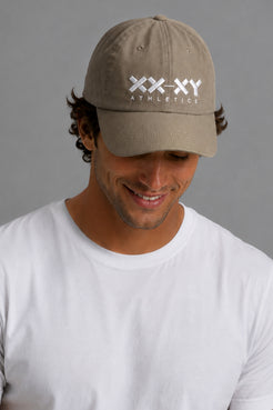 A man wearing a khaki baseball cap with a white 'XX-XY ATHLETICS' logo on the front, paired with a white t-shirt, photographed against a neutral gray background. || Mocha