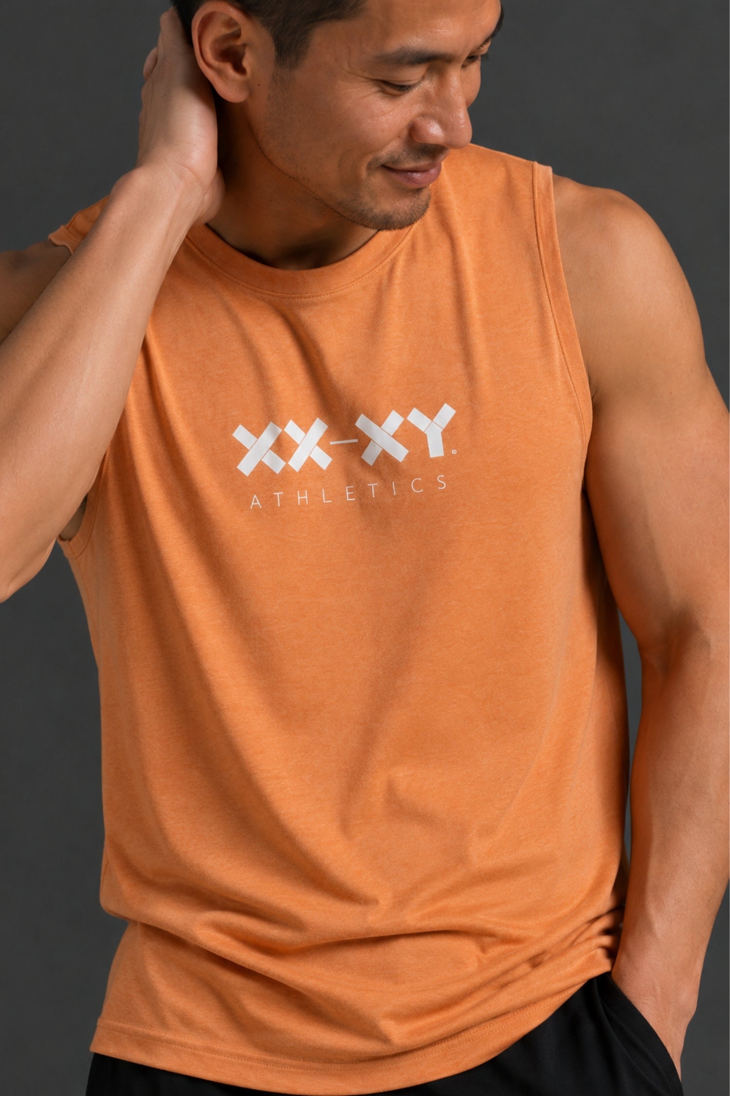 A man wearing an orange athletic tank top with a white 'XX-XY ATHLETICS' logo on the chest, photographed against a dark gray studio background.