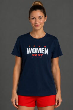A woman modeling a navy blue short-sleeve t-shirt with 'TEAM WOMEN XX XY FOOTBALL' printed in red and white on the front, paired with red athletic shorts, against a solid dark gray studio background. || Navy