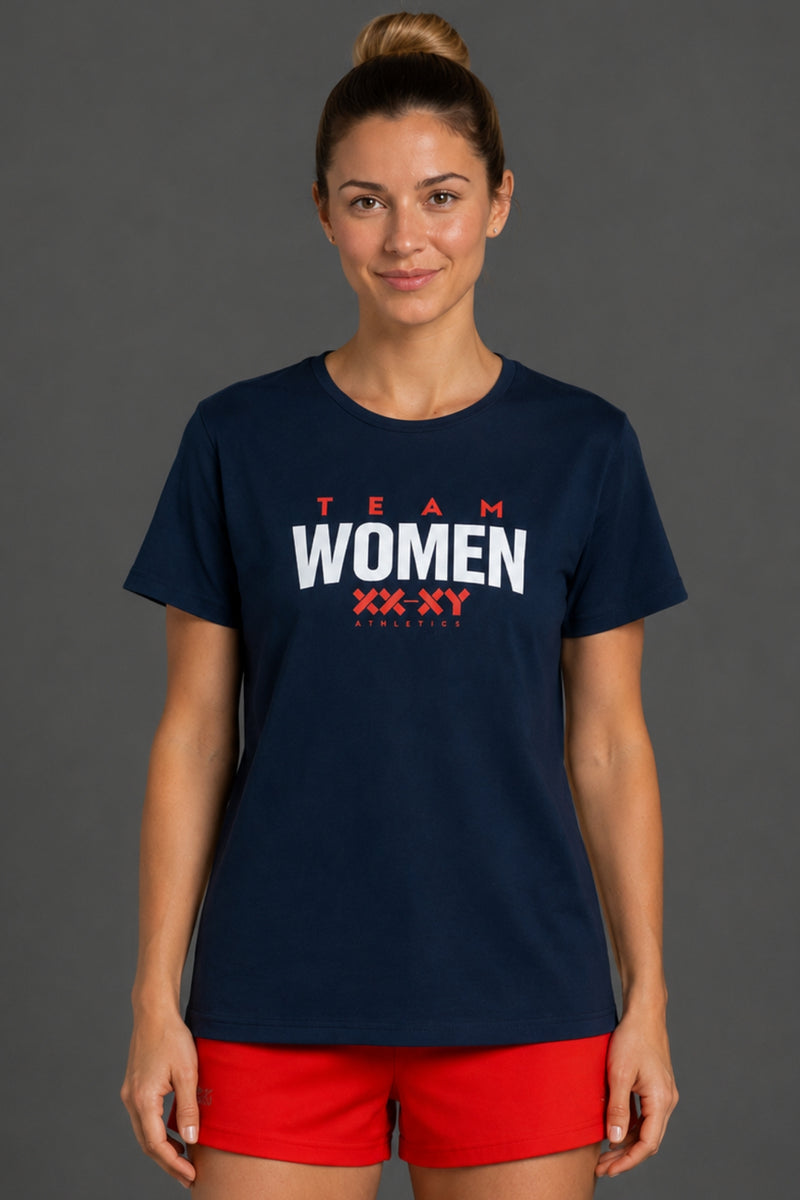 A woman modeling a navy blue short-sleeve t-shirt with 'TEAM WOMEN XX XY FOOTBALL' printed in red and white on the front, paired with red athletic shorts, against a solid dark gray studio background. || Navy