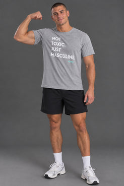 Male model wearing a gray “Not Toxic. Just Masculine.” graphic T-shirt with black shorts against a gray studio background. || Light Heather Gray