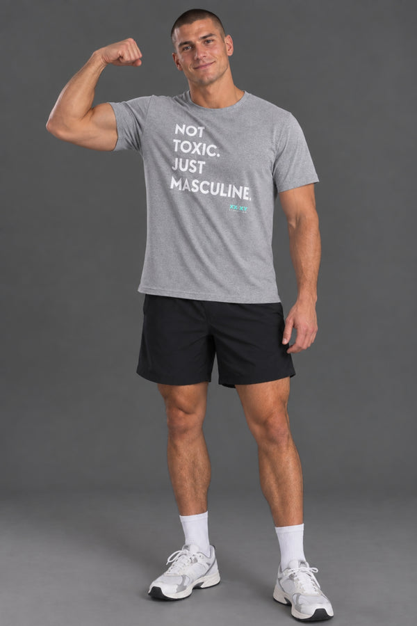 Male model wearing a gray “Not Toxic. Just Masculine.” graphic T-shirt with black shorts against a gray studio background. || Light Heather Gray