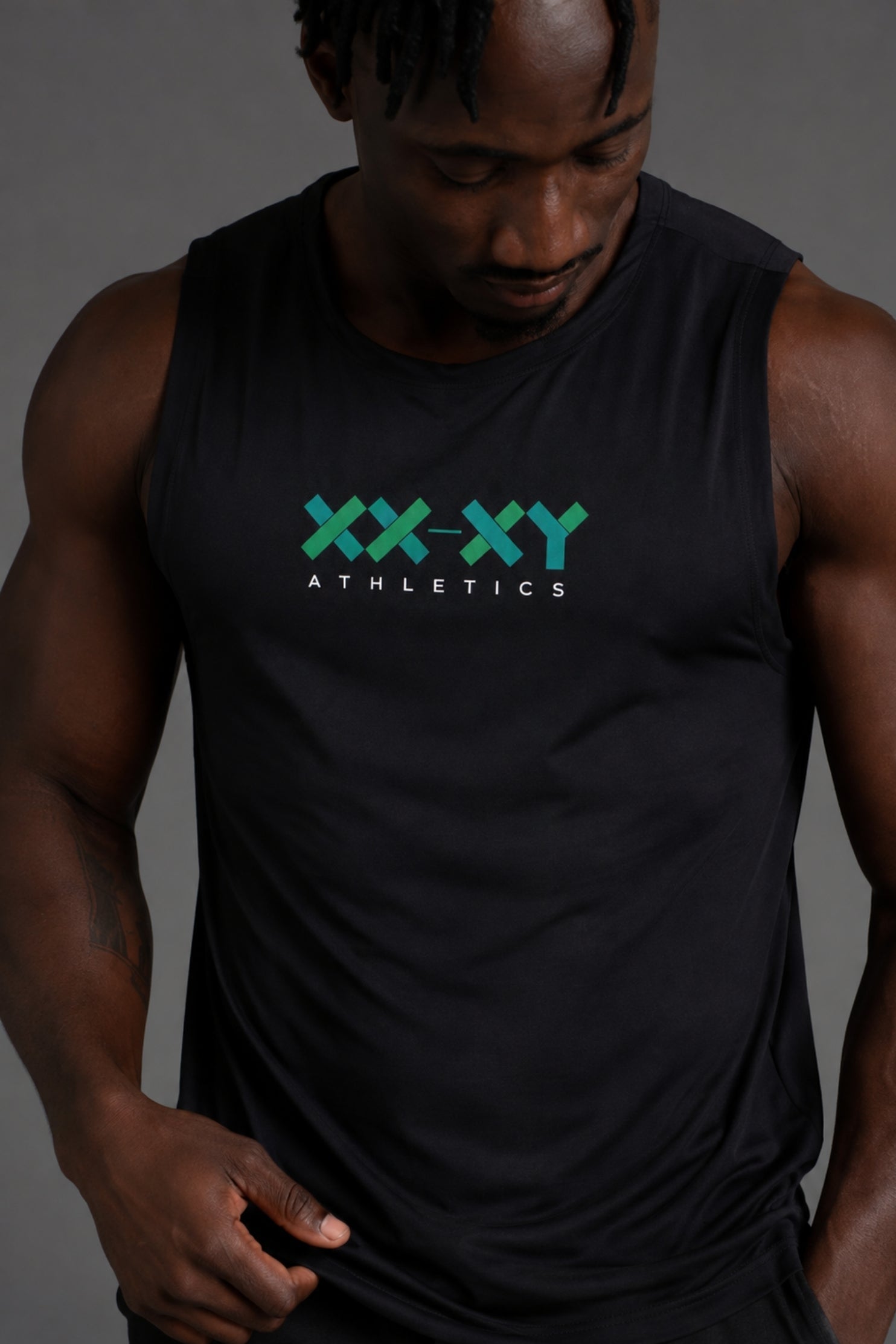 A man wearing a black sleeveless athletic tank top with a green and white 'XX-XY ATHLETICS' logo printed on the chest, photographed against a dark gray studio background. || Black
