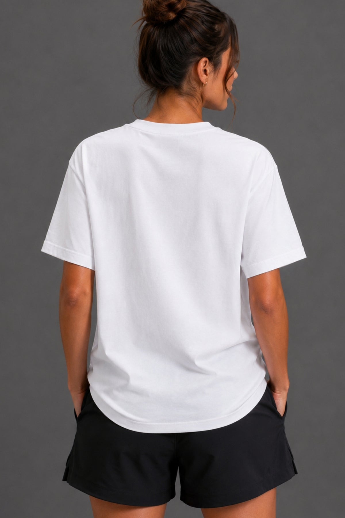 A woman viewed from behind wearing a plain white short-sleeve crewneck T-shirt and black shorts against a dark gray studio background.