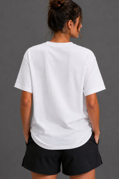 A woman viewed from behind wearing a plain white short-sleeve crewneck T-shirt and black shorts against a dark gray studio background. || White