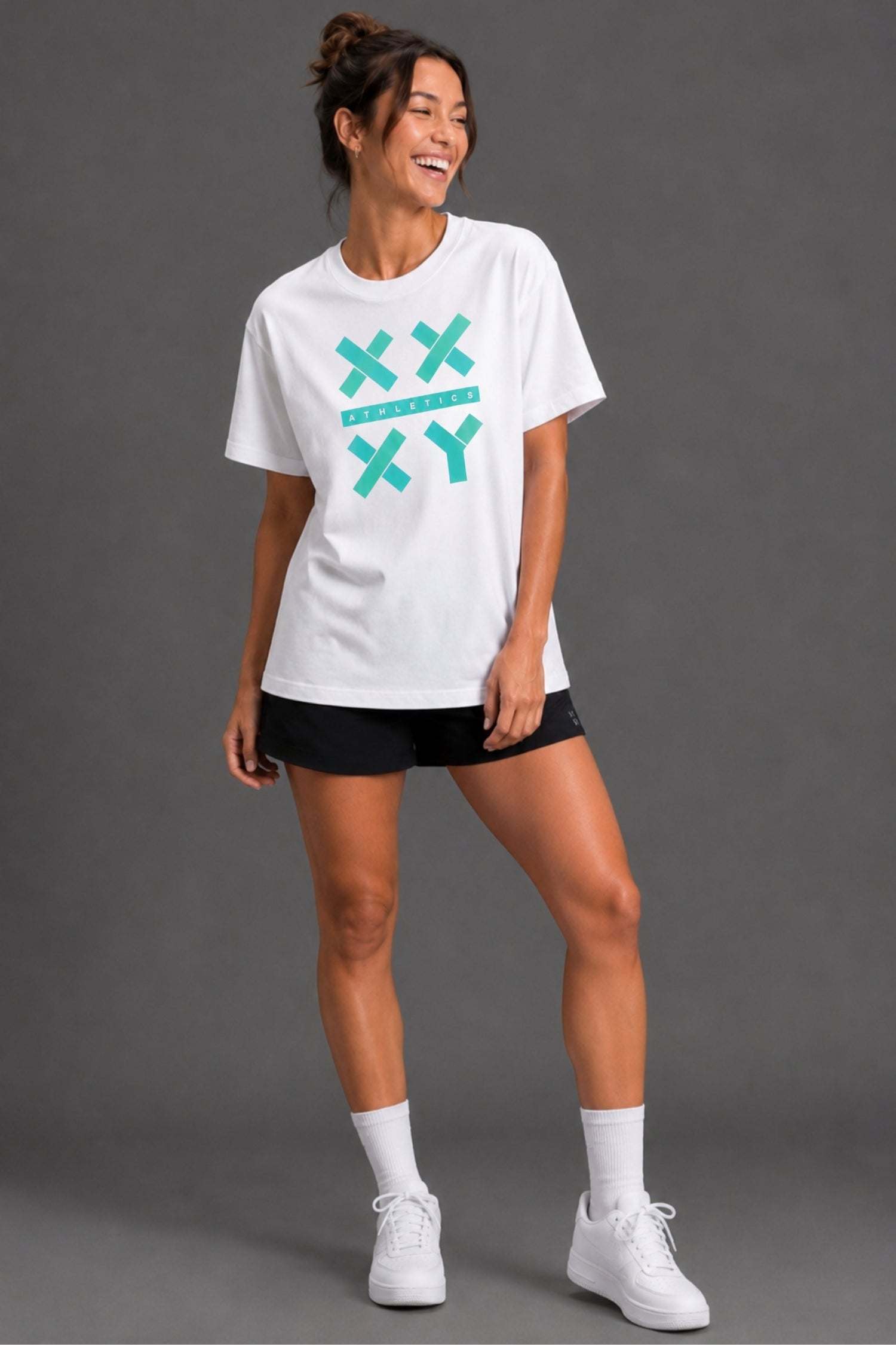 A woman modeling a white short-sleeve t-shirt with a teal graphic on the front, paired with black athletic shorts, white socks, and white sneakers, set against a solid gray studio background. || White