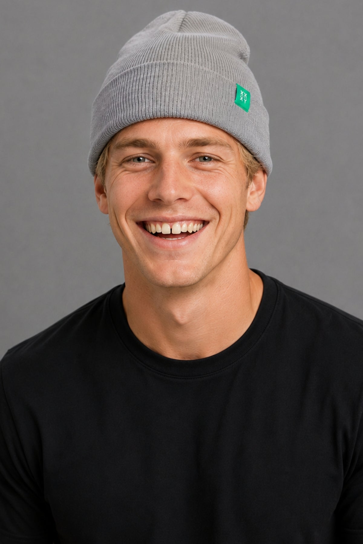 A smiling man wearing a light gray ribbed knit beanie with a small green square logo patch on the side, paired with a black crewneck shirt, against a solid gray studio background.