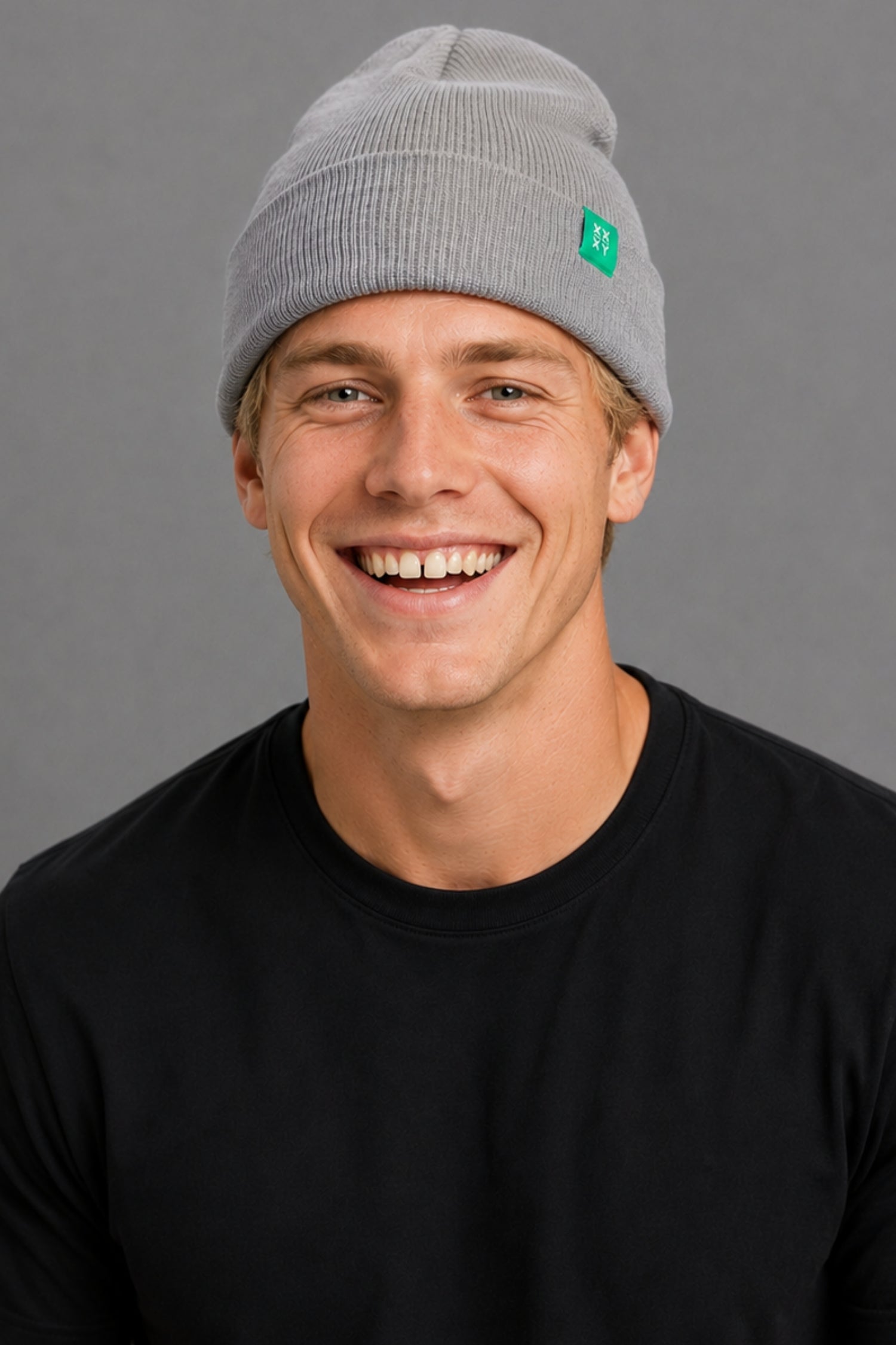 A smiling man wearing a light gray ribbed knit beanie with a small green square logo patch on the side, paired with a black crewneck shirt, against a solid gray studio background. || Light Heather Gray