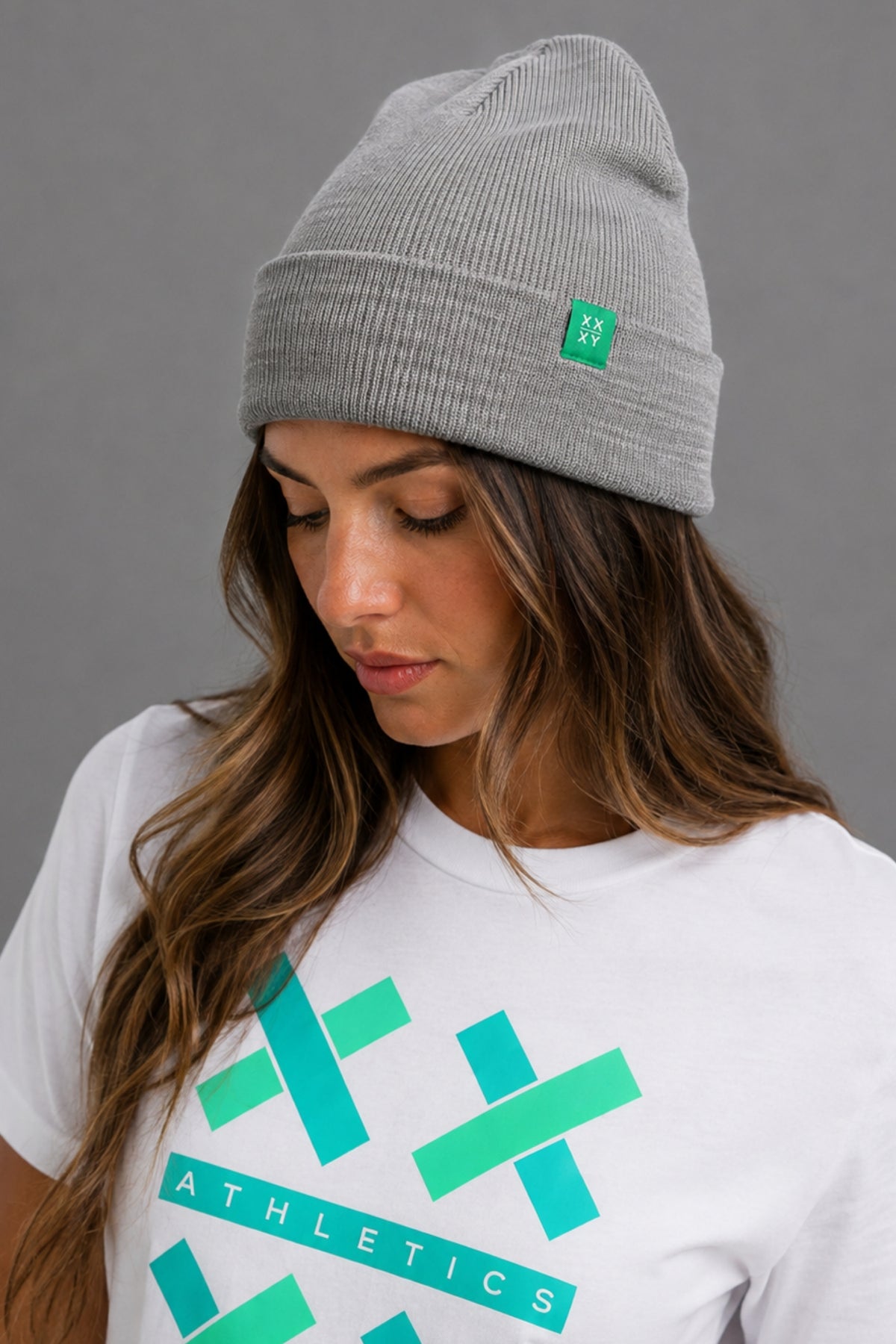 A woman wearing a light gray ribbed knit beanie with a small green square logo patch and a white t-shirt with a green athletic graphic, photographed against a solid gray studio background. || Light Heather Gray