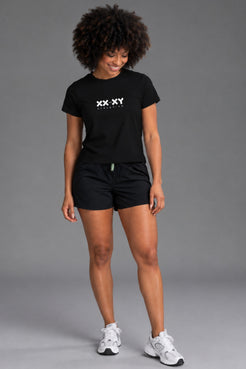 Woman wearing a black XX-XY Athletics logo tee with black athletic shorts against a gray studio background. || Black
