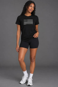 Female model wearing a black XX-XY Athletics American flag graphic tee, black athletic shorts, white socks, and sneakers against a gray background || Black