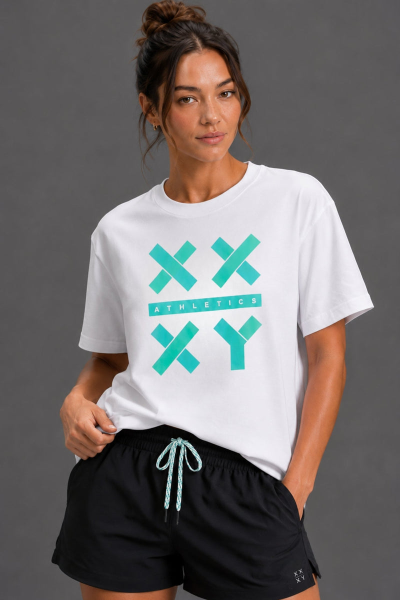 A woman wearing a white crewneck t-shirt with a teal 'XX ATHLETICS XY' graphic on the front, paired with black athletic shorts, against a solid dark gray studio background. || White