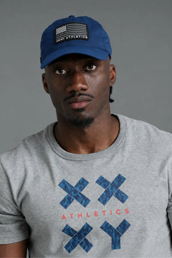 Black male model wearing a blue XX-XY Athletics American flag hat and gray graphic T-shirt against a gray studio background. || Blue